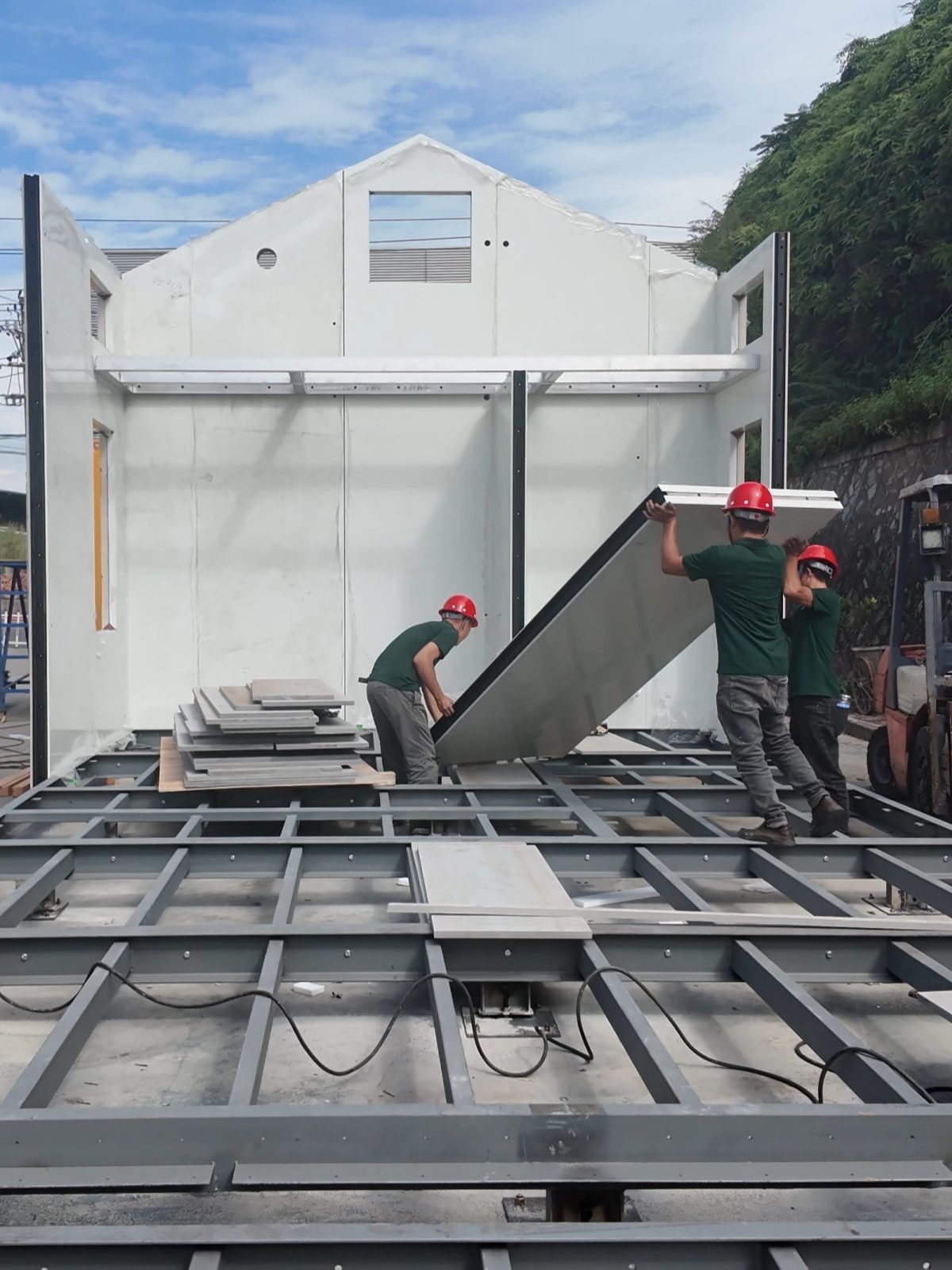 Two workers lifting a prefabricated gable wall panel onto the steel floor frame